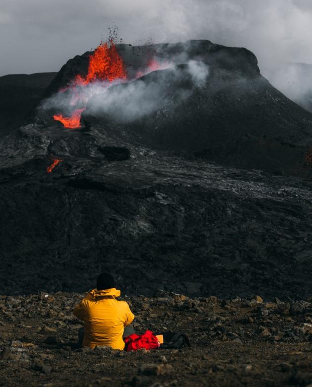 A person in a yellow winter jacket sits in front of an erupting volcano